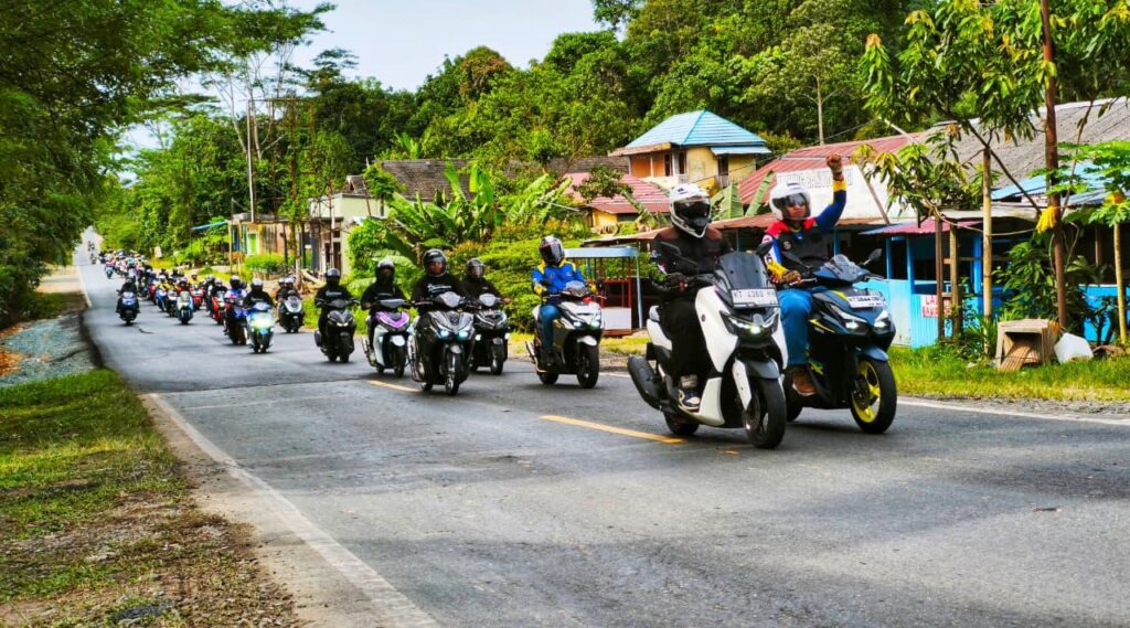 Ratusan riders Yamaha tampak konvoi santai melintasi jalan poros dengan latar perkampungan dan pepohonan hijau saat agenda SUNMORYGAB jelang Ramadan.